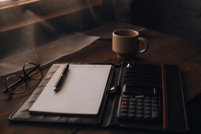 Dnevnik, kalkulator i šalica kave na drvenom stolu, uz olovku i naočale.